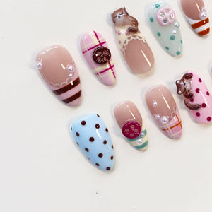 A set of almond-shaped press-on nails featuring various designs including pink and white plaid patterns, light blue and mint green polka dots, brown button embellishments, small white pearls, and miniature brown cat figures.