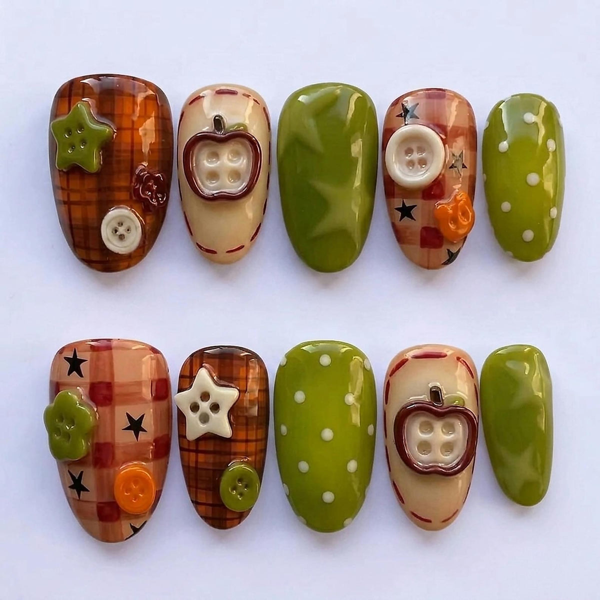 A set of ten press-on nails featuring a variety of designs including brown plaid patterns, olive green surfaces with white polka dots, and three-dimensional embellishments such as small plastic buttons, stars, and apple-shaped charms.