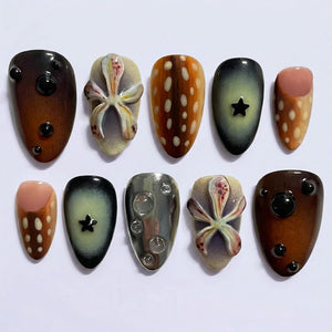 A set of ten press-on nails featuring diverse designs including brown gradient surfaces with black raised spheres, textured floral-like relief patterns, brown and white spotted motifs, dark green gradients with black stars, and metallic silver with c