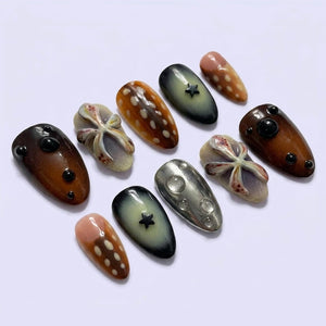 A set of ten press-on nails featuring varied designs including brown with white spots, dark grey with black stars, metallic silver with clear bubbles, and light-colored nails with raised, textured, flower-like sculptural elements.