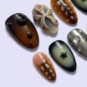 A collection of six almond-shaped press-on nails featuring diverse designs. One is dark brown with black raised dots, another has a beige bow, one is amber with white spots, one is metallic silver, one is black with a star, and one is peach.