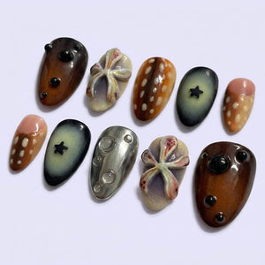 A set of ten press-on nails featuring diverse designs including brown surfaces with black spheres, beige bases with raised floral-like shapes, brown surfaces with white dots, dark grey surfaces with black stars, and a metallic silver finish.