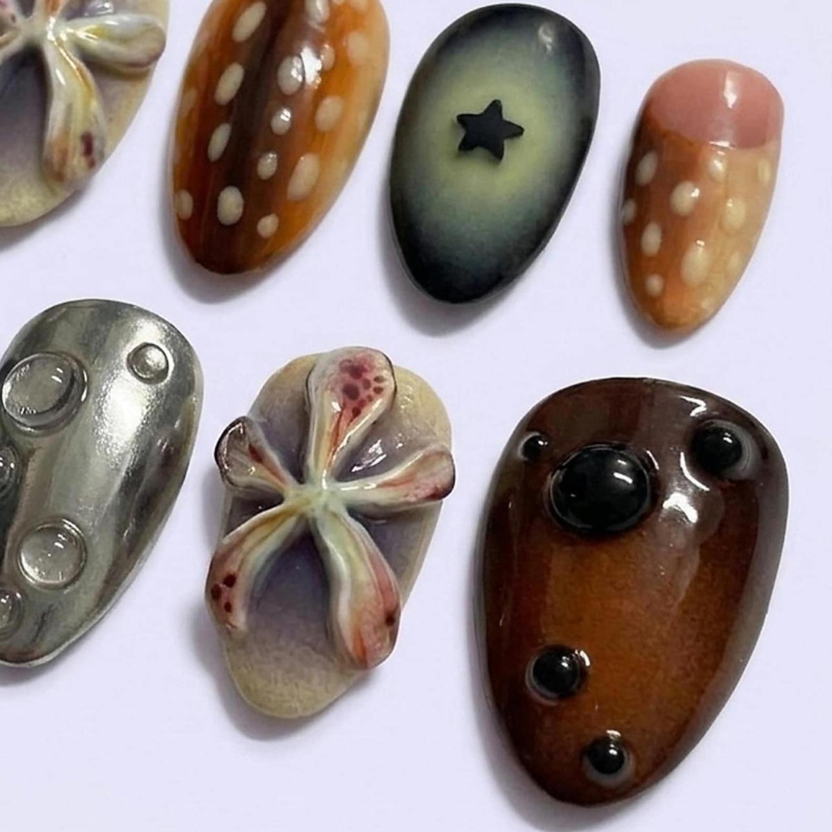 A collection of seven decorative nail art pieces featuring diverse designs including a brown spotted pattern, a dark grey piece with a black star, a pink and brown spotted piece, a metallic silver piece with bubbles, and floral motifs.