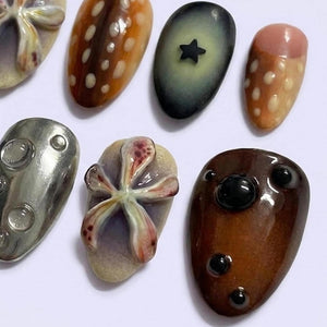 A collection of seven decorative nail art pieces featuring diverse designs including a brown spotted pattern, a dark grey piece with a black star, a pink and brown spotted piece, a metallic silver piece with bubbles, and floral motifs.