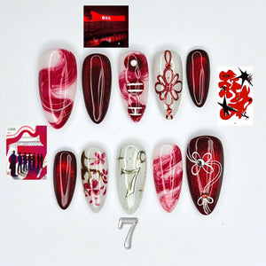 A set of ten almond-shaped press-on nails featuring a red and white color palette. Designs include red marble swirls, deep red glitter, a red decorative knot, a cherry blossom branch, a silver number seven, and small rhinestones.