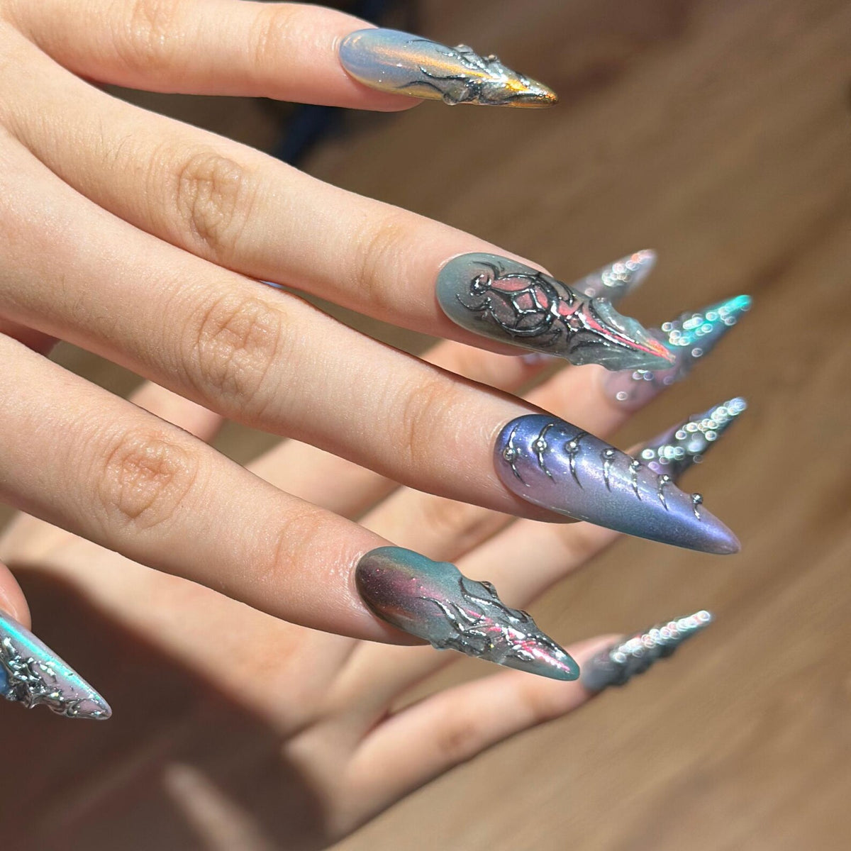 Long stiletto-shaped artificial nails featuring a multi-tonal iridescent finish in shades of blue, purple, and grey. The nails are decorated with raised silver metallic accents, intricate filigree-like patterns, and small embedded crystals.