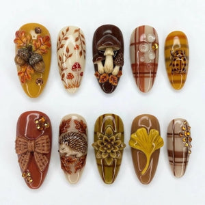 A set of ten almond-shaped press-on nails featuring autumn-themed designs including 3D acorns, mushrooms, a hedgehog, a bow, a ginkgo leaf, plaid patterns, and small amber-colored rhinestones in shades of brown, orange, and cream.