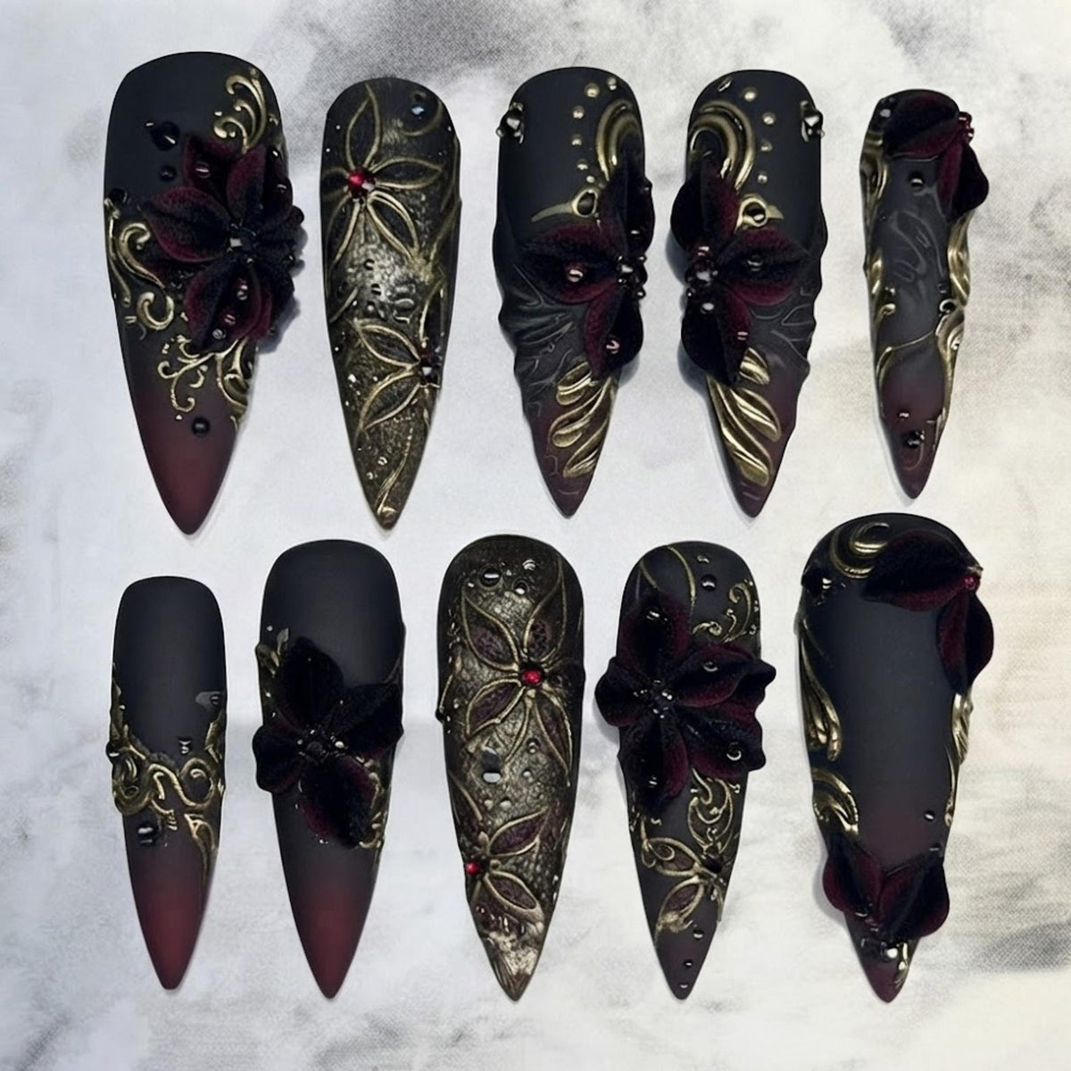 A set of ten stiletto-shaped press-on nails featuring a matte black and deep burgundy gradient base. The nails are decorated with raised gold filigree patterns, three-dimensional dark red velvet flowers, and small red rhinestones.