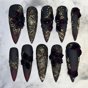 A set of ten stiletto-shaped press-on nails featuring a matte black and deep burgundy gradient base. The nails are decorated with raised gold filigree patterns, three-dimensional dark red velvet flowers, and small red rhinestones.