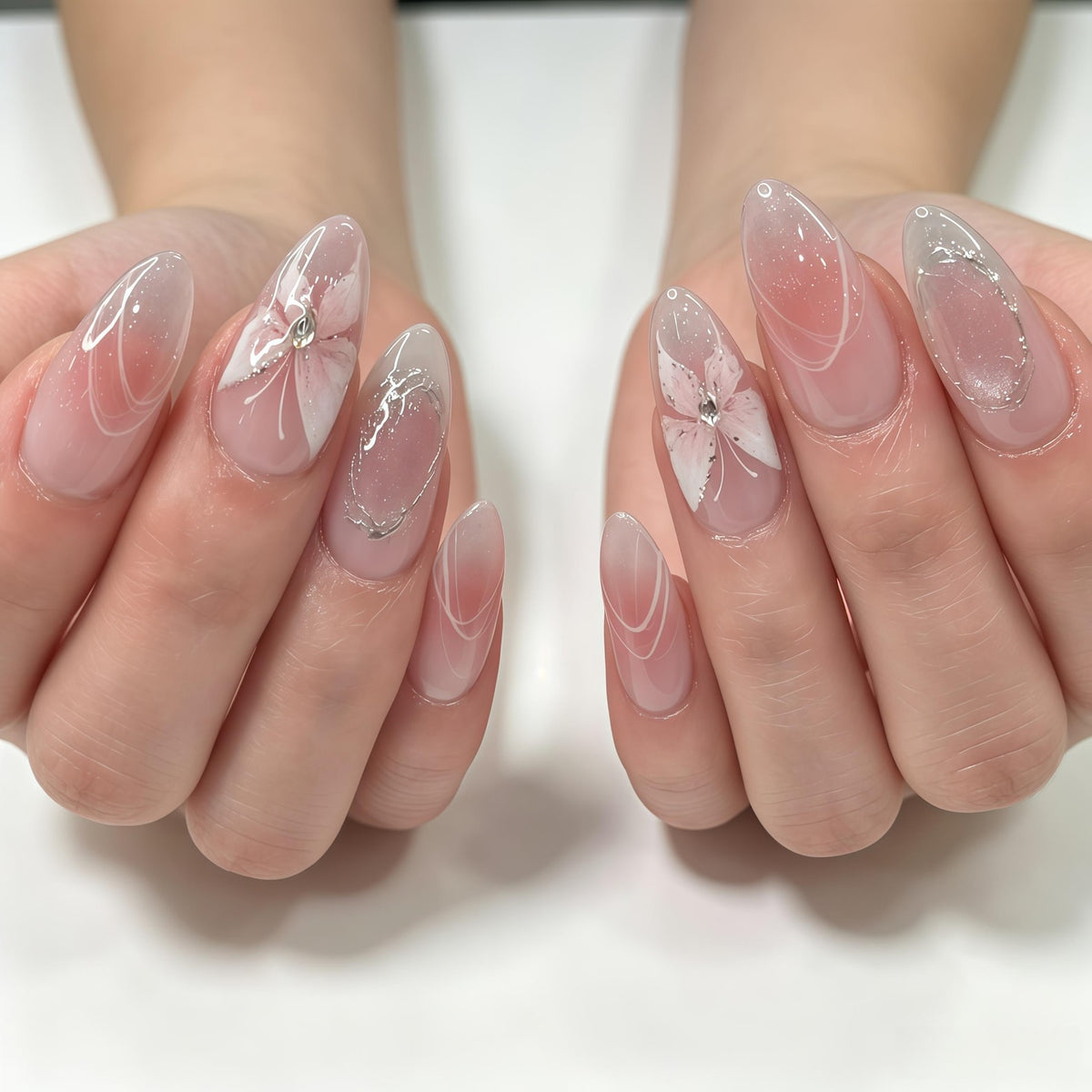 Almond-shaped nails featuring a sheer pink base with a soft gradient effect. The design includes delicate white hand-painted floral petals, silver metallic line accents, and small clear rhinestones placed at the center of the flowers.