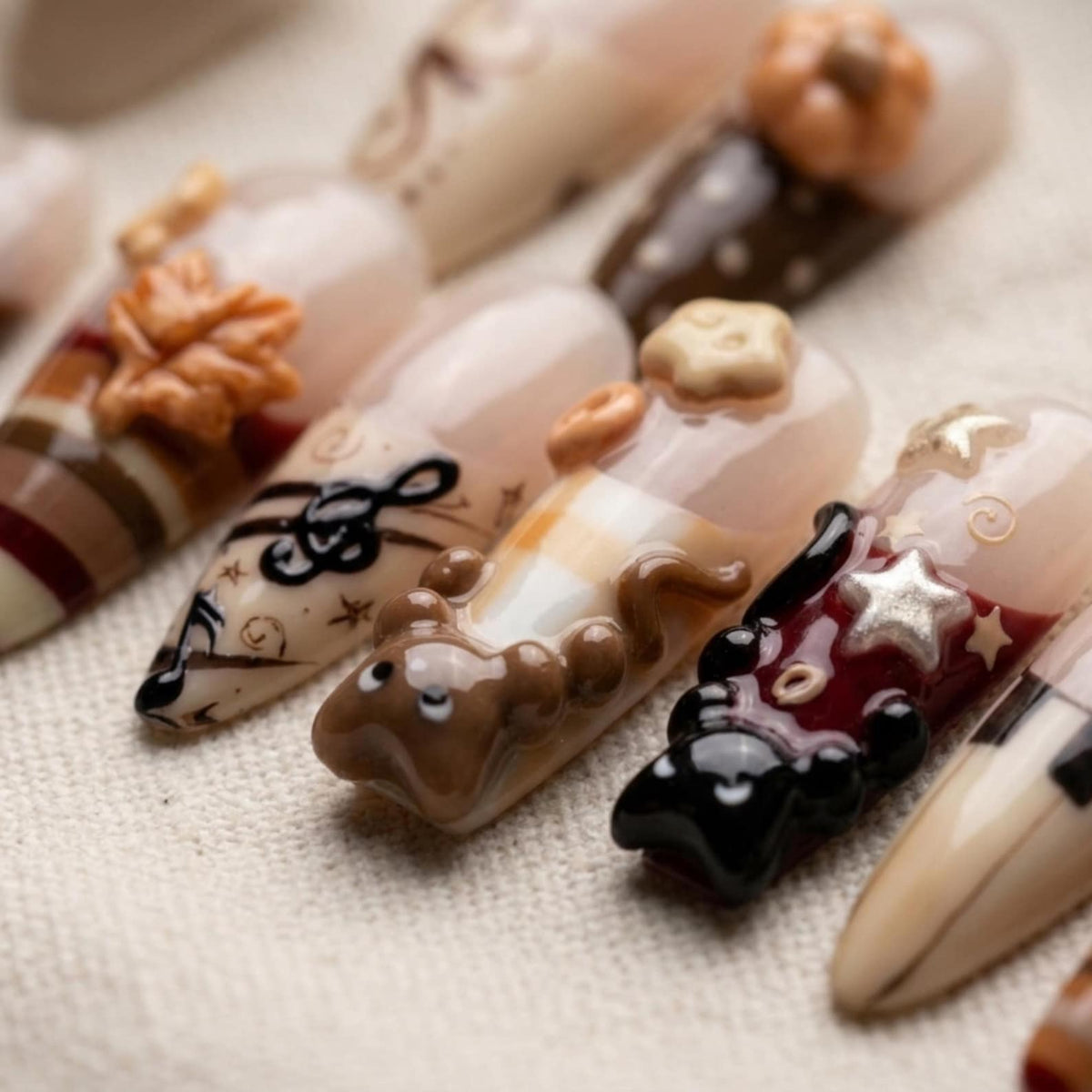 A set of press-on nails featuring various autumn-themed designs including 3D sculpted elements like a brown leaf, a pumpkin, a small brown animal, and a star. The nails display patterns of stripes, musical notes, and solid colors.