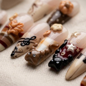A set of press-on nails featuring various autumn-themed designs including 3D sculpted elements like a brown leaf, a pumpkin, a small brown animal, and a star. The nails display patterns of stripes, musical notes, and solid colors.