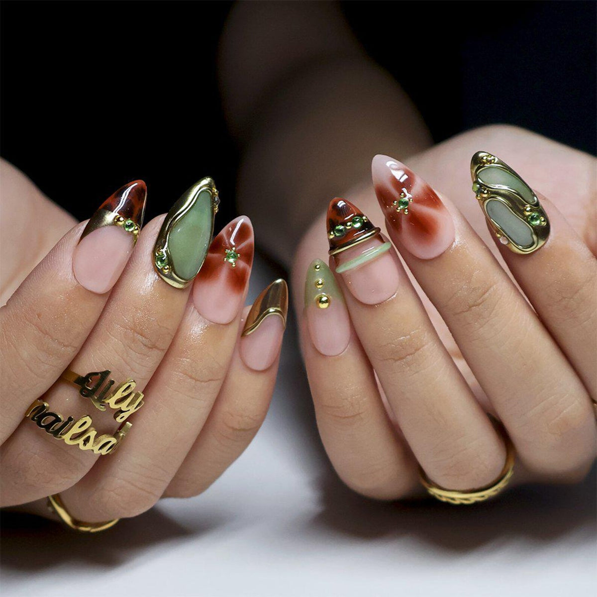 Two hands held together showing medium almond nails: sheer nude bases with brown tortoiseshell tips, sage green stone-look panels rimmed in gold, gold chrome swirls, and tiny green gems. Tips extend about a centimetre past the fingertips.