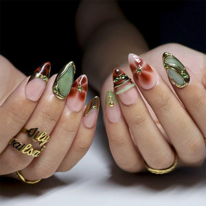 Two hands held together showing medium almond nails: sheer nude bases with brown tortoiseshell tips, sage green stone-look panels rimmed in gold, gold chrome swirls, and tiny green gems. Tips extend about a centimetre past the fingertips.