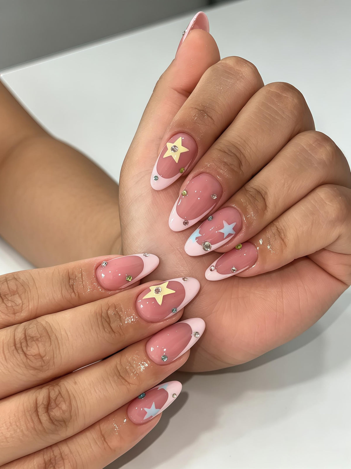 Almond-shaped nails featuring a glossy nude base with light pink French tips. The design includes small pastel yellow and light blue star-shaped decals on select nails, accented with tiny, scattered clear rhinestones throughout.