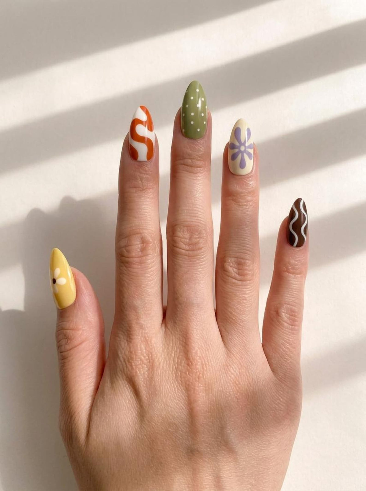 Almond-shaped press-on nails featuring a variety of designs including a yellow daisy, orange and white swirls, white polka dots on olive green, a purple flower on cream, and white wavy lines on a dark brown base.