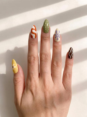 Almond-shaped press-on nails featuring a variety of designs including a yellow daisy, orange and white swirls, white polka dots on olive green, a purple flower on cream, and white wavy lines on a dark brown base.