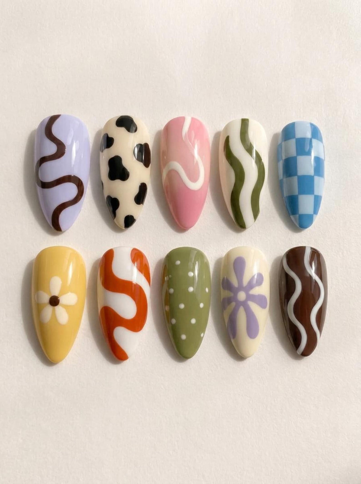 Ten almond-shaped press-on nails featuring various designs: a lavender nail with a brown squiggle, a cream nail with black cow spots, a pink nail with a white swirl, a cream nail with green waves, and a blue and white checkered nail.