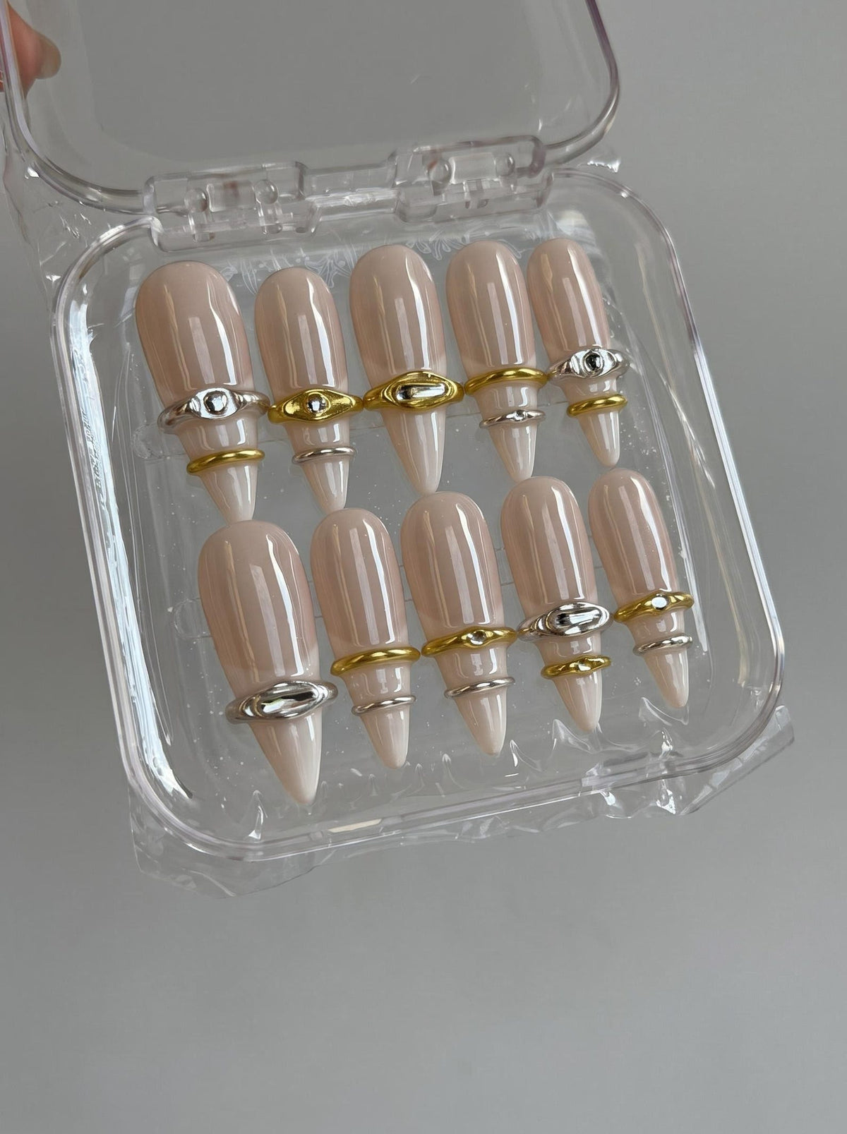 A set of ten almond-shaped press-on nails in a glossy nude beige finish, each adorned with a single metallic ring accent in either gold or silver tones, presented in a clear plastic storage container with a hinged lid.