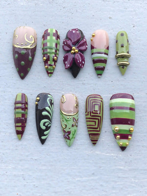 A set of ten almond-shaped press-on nails featuring a color palette of burgundy, light green, and nude. The designs include plaid patterns, a three-dimensional purple flower, gold metallic studs, swirling filigree, and geometric lines.