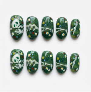 A set of ten press-on nails featuring a dark green base with a glittery finish. The design includes white skeletal hand and arm bones, small white and yellow flowers, and tiny red and blue dots scattered across the surface of each nail.