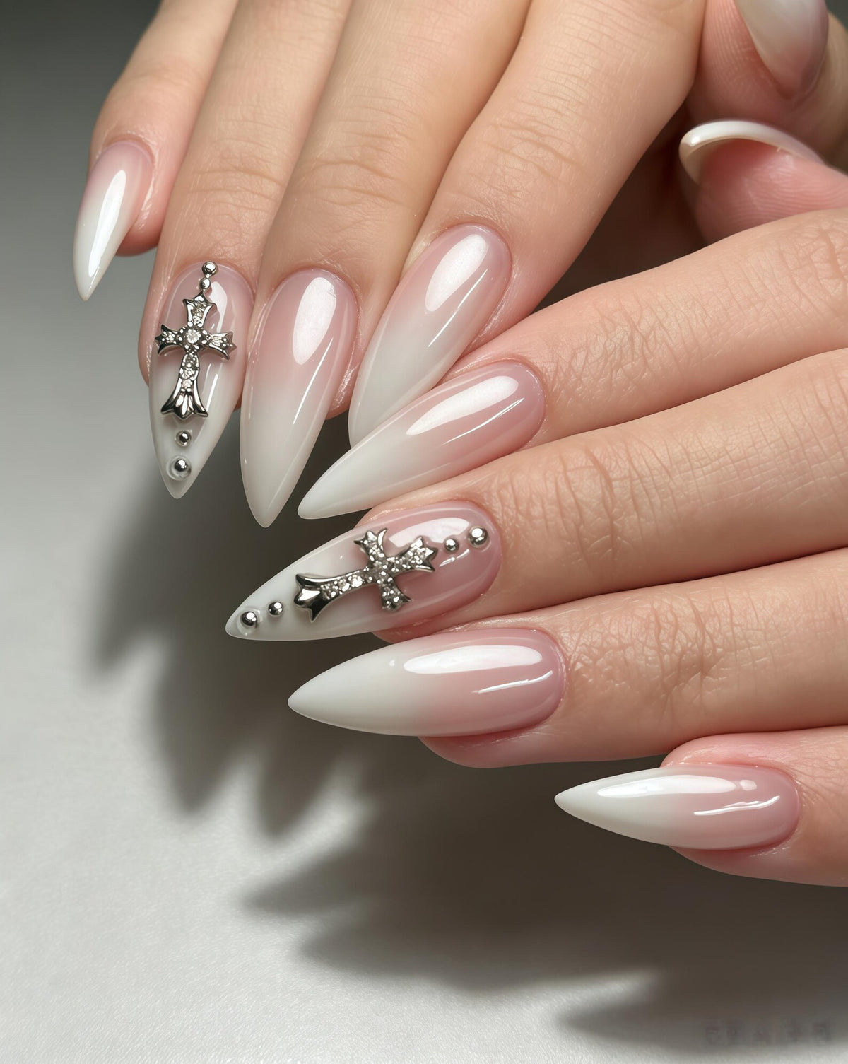 Long, pointed stiletto-shaped nails featuring a soft pink to white ombre gradient. Two of the nails are decorated with silver-toned metal cross charms encrusted with small clear crystals, accented by tiny silver spherical studs.
