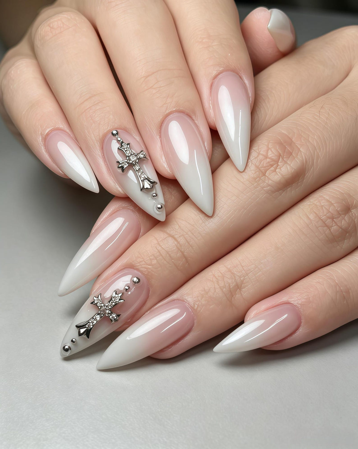 Long, pointed stiletto-shaped nails featuring a soft white and nude pink ombre gradient. Two nails are accented with silver-toned metal cross charms embedded with small clear crystals, complemented by tiny silver metallic spheres.