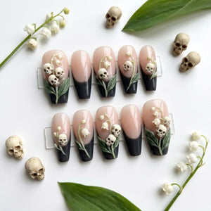 A set of ten press-on nails featuring a nude base with black French tips. Each nail is adorned with three-dimensional white lily of the valley flowers and small beige skulls. The set is displayed alongside loose miniature skulls and lily of the valle