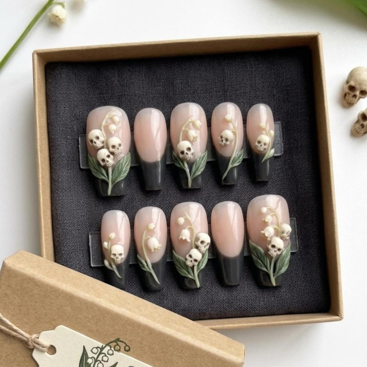 A set of ten press-on nails featuring a French manicure design with a dark gray tip and a nude base. Several nails are decorated with three-dimensional white skull accents and green lily of the valley floral embellishments.
