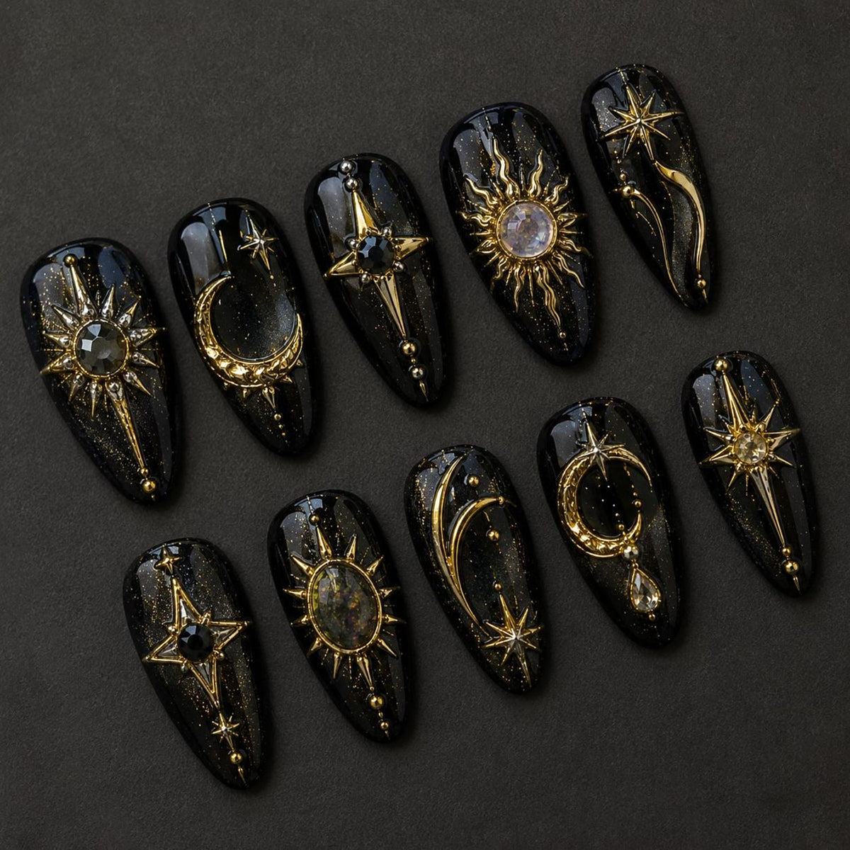 A set of ten almond-shaped press-on nails with a black base and gold glitter finish. Each nail features intricate gold metal charms including sunbursts, crescent moons, and stars, accented with small clear and black rhinestones.
