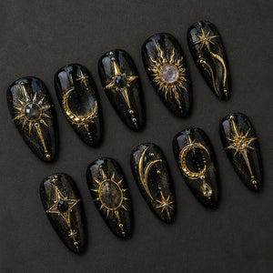 A set of ten almond-shaped press-on nails with a black base and gold glitter finish. Each nail features intricate gold metal charms including sunbursts, crescent moons, and stars, accented with small clear and black rhinestones.