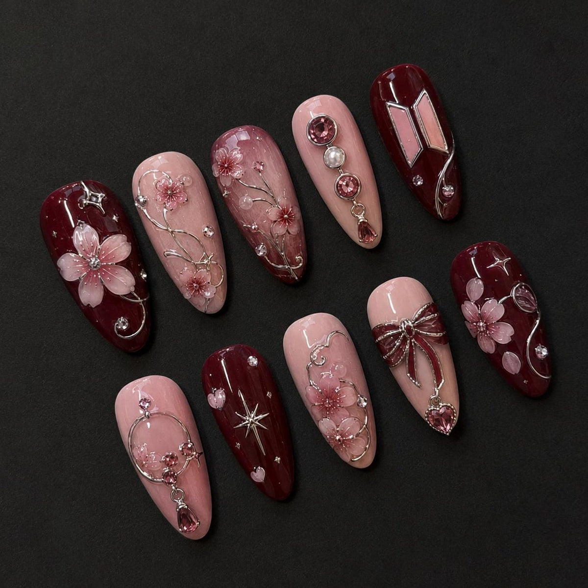 A set of ten almond-shaped press-on nails featuring a color palette of deep burgundy and soft blush pink. The designs include 3D cherry blossom motifs, silver metallic accents, dangling heart charms, starbursts, and pearl embellishments.