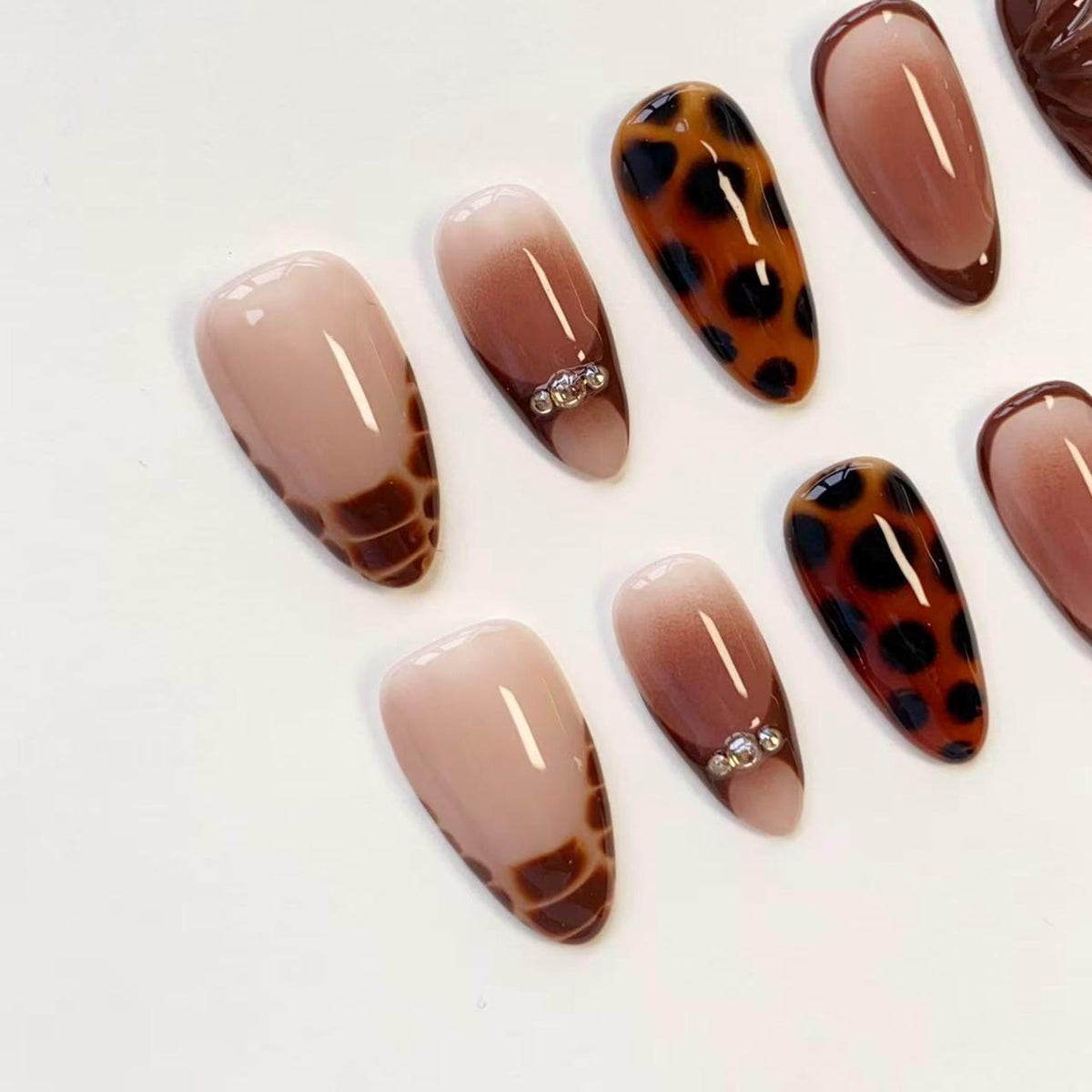 A set of almond-shaped press-on nails featuring a variety of designs including brown and tan crocodile skin patterns, leopard print spots, and solid nude gradients, with some nails accented by small, clear, round decorative rhinestones.