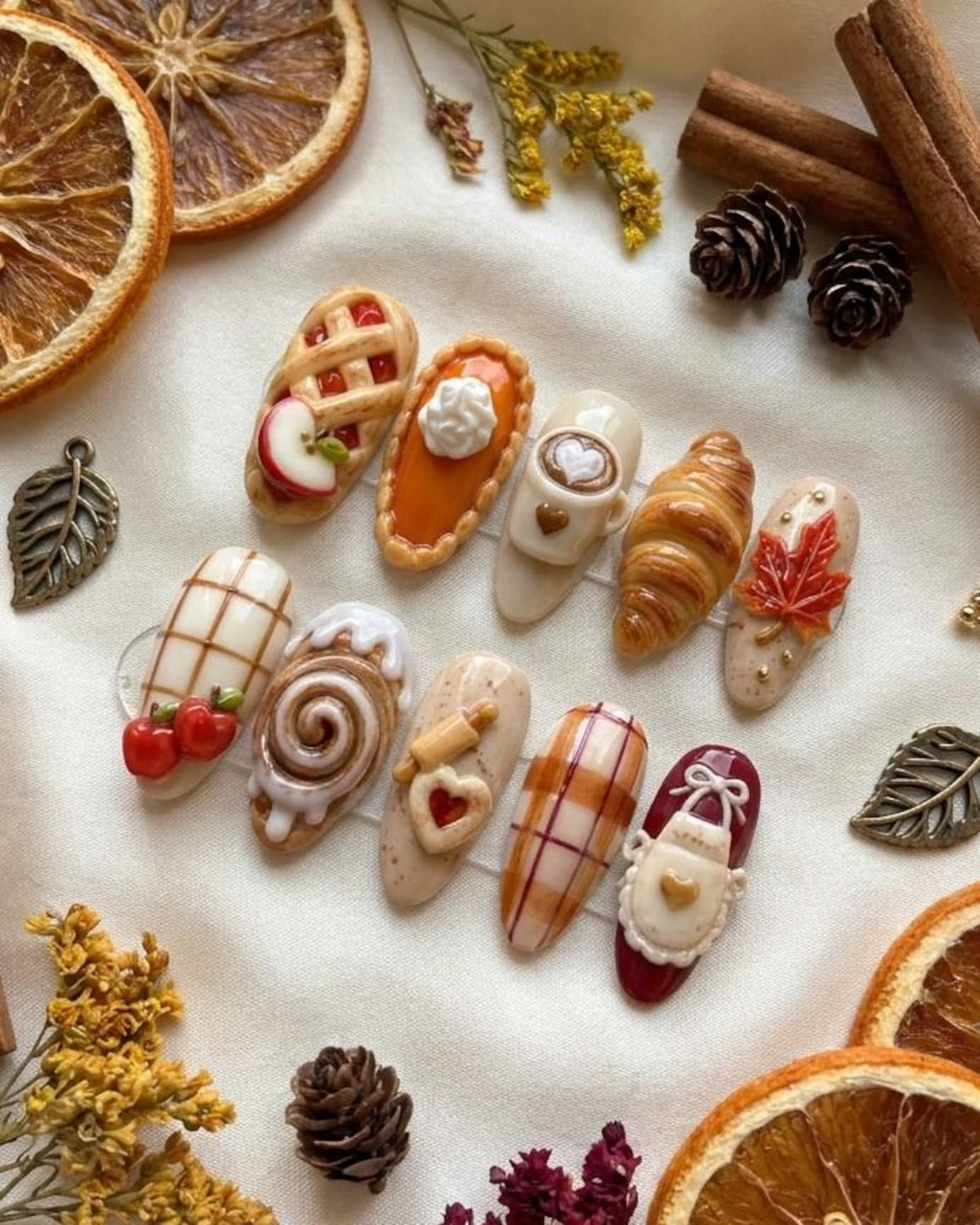 A set of ten press-on nails featuring autumn-themed 3D designs including an apple pie, pumpkin pie, latte, croissant, maple leaf, plaid patterns, cinnamon roll, rolling pin with heart, and a white apron with a heart on a maroon base.