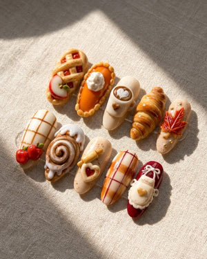 A set of ten press-on nails featuring 3D miniature food and autumn-themed designs, including an apple pie, pumpkin pie, latte, croissant, maple leaf, cinnamon roll, rolling pin, plaid patterns, and a white apron with a heart.