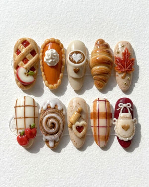 A set of ten press-on nails featuring 3D autumn-themed designs including an apple pie, pumpkin pie, latte art, croissant, maple leaf, plaid patterns, cinnamon roll, rolling pin with heart cookie, and a white apron on a dark red base.