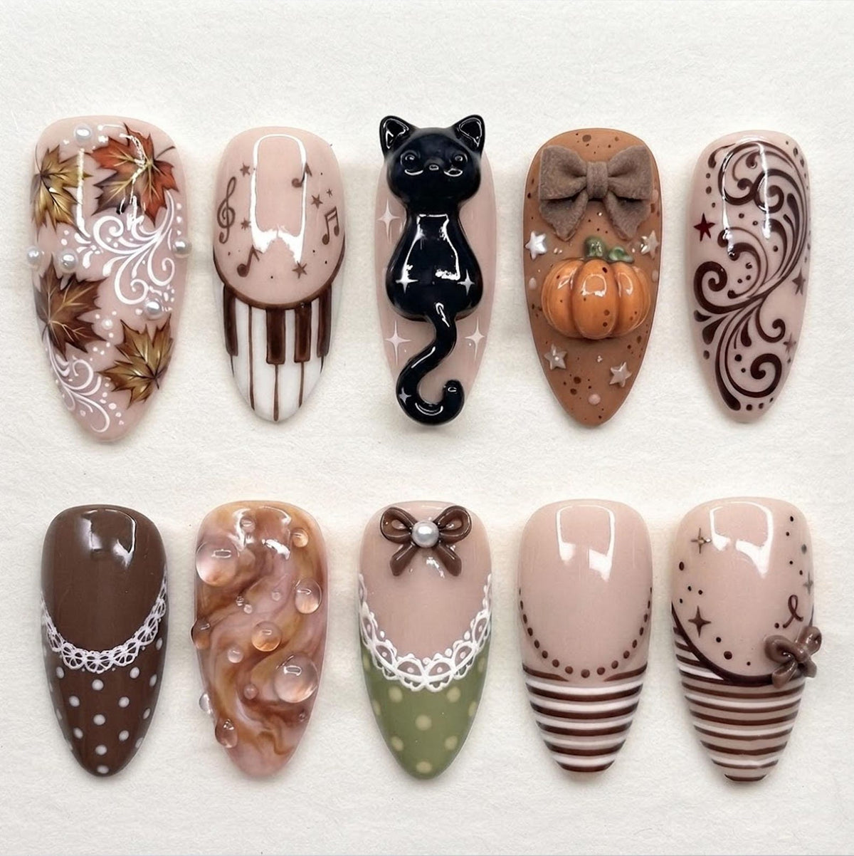 A set of ten press-on nails featuring various designs including autumn leaves, piano keys, a 3D black cat, a 3D pumpkin with a bow, brown swirls, white polka dots, lace patterns, stripes, and small bows on a neutral beige base.