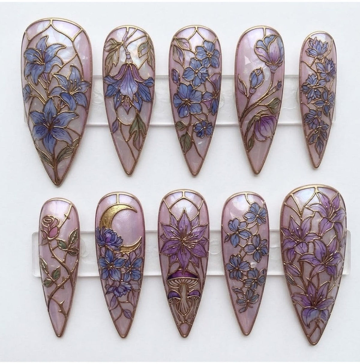 A set of ten stiletto-shaped press-on nails featuring a light pink base with a stained-glass effect. Each nail displays intricate gold-lined illustrations of purple and blue flowers, vines, a crescent moon, and mushrooms.