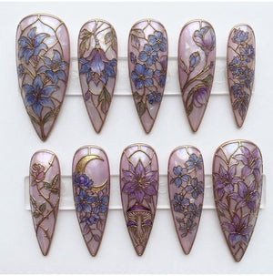 A set of ten stiletto-shaped press-on nails featuring a light pink base with a stained-glass effect. Each nail displays intricate gold-lined illustrations of purple and blue flowers, vines, a crescent moon, and mushrooms.