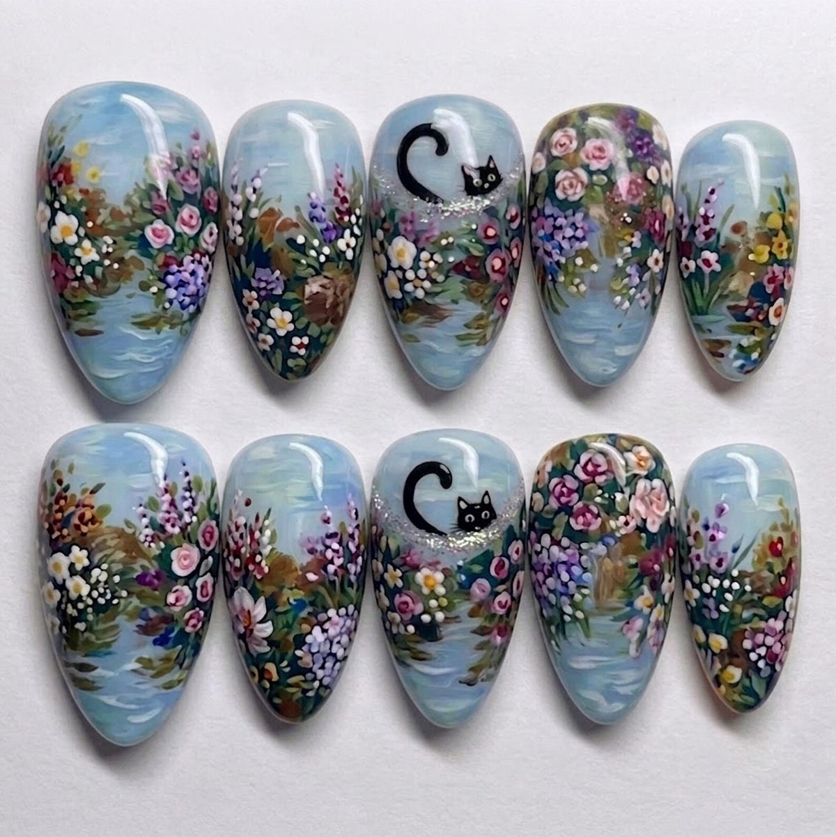 A set of ten almond-shaped press-on nails featuring a hand-painted garden scene with a light blue background. The design includes colorful floral clusters in pink, purple, and white, with a small black cat peeking out on two nails.