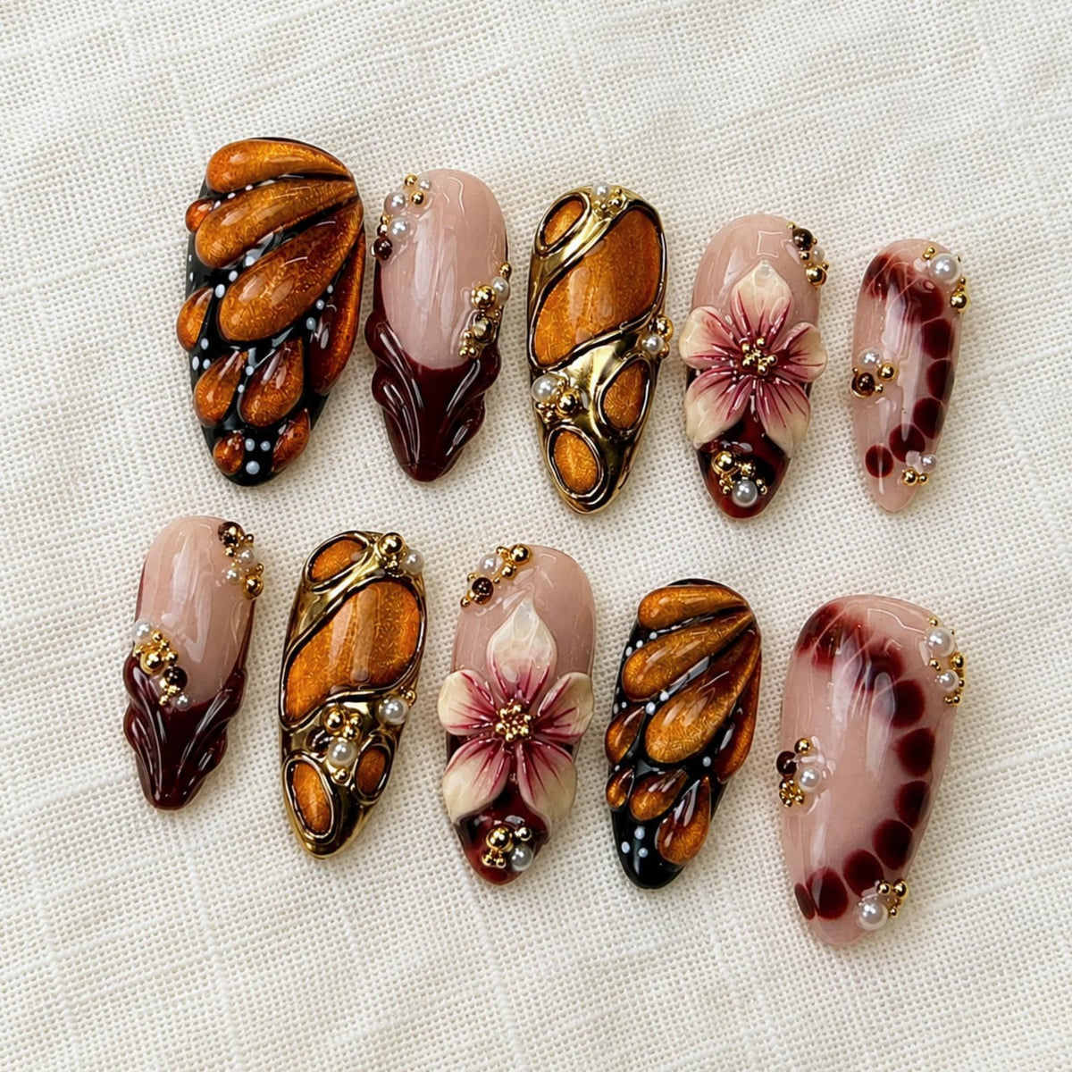 A set of ten press-on nails featuring a variety of designs including amber-colored metallic shapes, dark maroon accents, sculpted floral motifs, and small gold and white beads arranged on a nude base for a decorative manicure set.