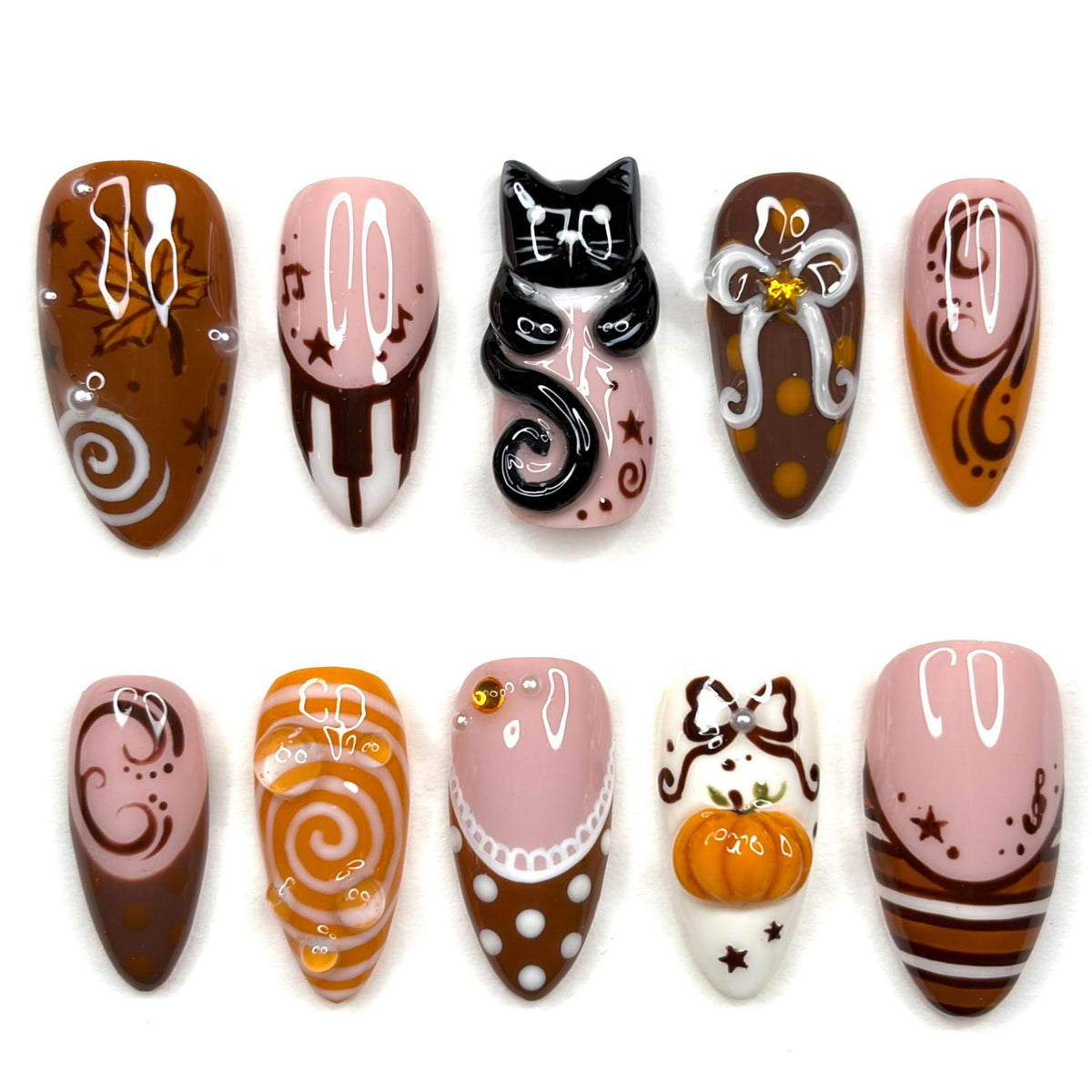 A set of ten press-on nails featuring autumn-themed designs including a black cat, a pumpkin, maple leaves, swirls, polka dots, and musical notes in a color palette of brown, orange, white, and pink with glossy finishes and 3D accents.