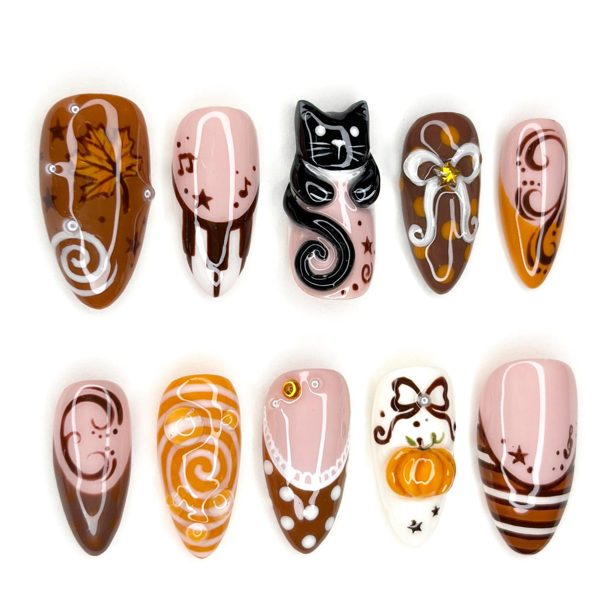 A set of ten press-on nails featuring autumn-themed designs including a brown maple leaf, a black cat figure, a small orange pumpkin, white bows, musical notes, stars, and swirling patterns in shades of brown, orange, white, and pink.