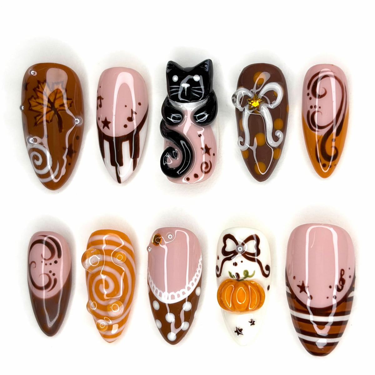 A set of ten press-on nails featuring autumn-themed designs including a black cat figure, a 3D orange pumpkin, brown and white swirls, maple leaves, bows, and musical notes on a palette of brown, white, and soft pink glossy finishes.