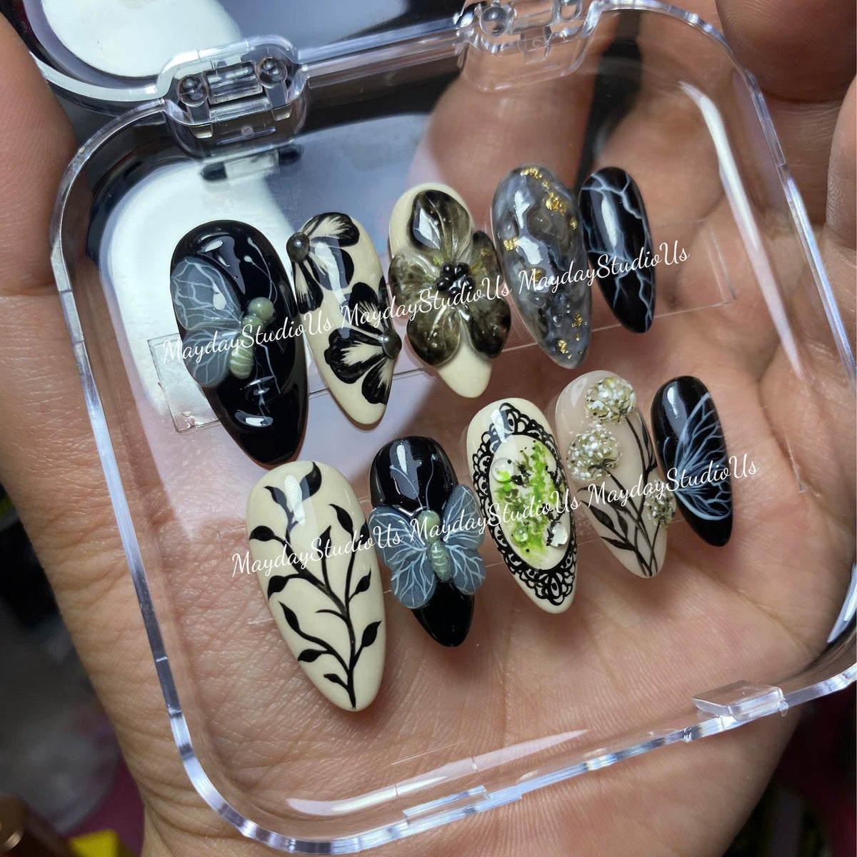 A set of ten almond-shaped press-on nails in a clear plastic case. The designs feature black and cream backgrounds with hand-painted floral motifs, butterfly accents, marble patterns with gold flakes, and small white pearl-like embellishments.