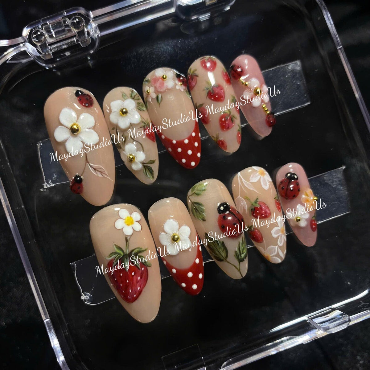 A set of ten almond-shaped press-on nails featuring a nude base with hand-painted designs including red strawberries, white flowers, red and white polka dots, and small ladybugs. The text MaydayStudioUs is visible on the nails.