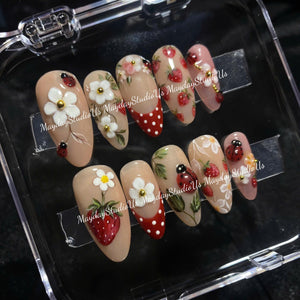A set of ten almond-shaped press-on nails featuring a nude base with hand-painted designs including red strawberries, white flowers, red and white polka dots, and small ladybugs. The text MaydayStudioUs is visible on the nails.