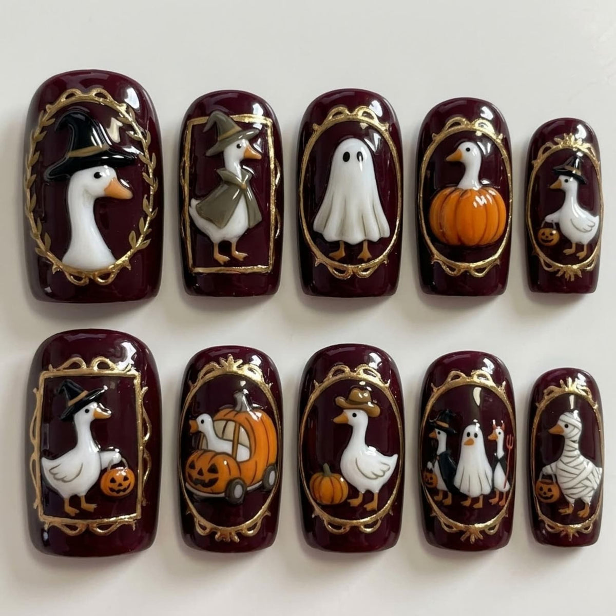 A set of ten press-on nails with a deep burgundy base, each featuring a raised 3D design framed in gold. The designs include white geese dressed in various Halloween costumes like a witch, ghost, mummy, and pumpkin-themed outfits.