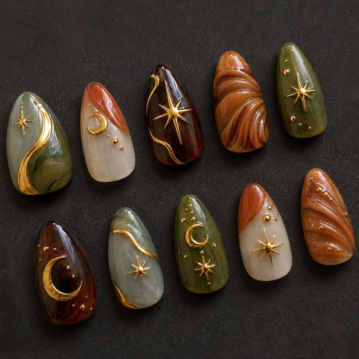 A set of ten almond-shaped press-on nails featuring a celestial theme. The designs include deep olive green, brown, and cream marbled patterns accented with gold-toned metal charms shaped like crescent moons and four-pointed stars.