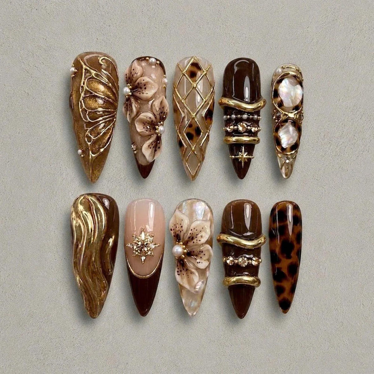 A set of ten almond-shaped press-on nails featuring brown, gold, and cream tones. Designs include gold filigree, 3D floral accents, leopard print, metallic swirls, pearl embellishments, and geometric gold lattice patterns on a glossy finish.